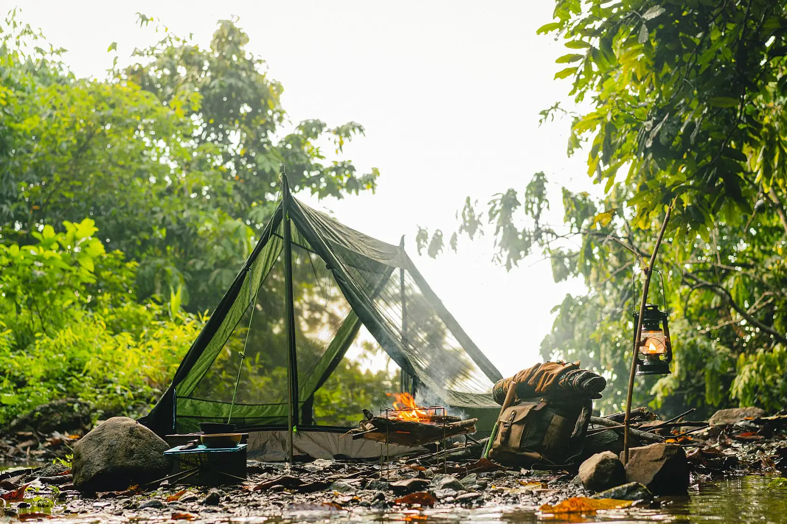 A serene camping site with a tent, campfire, and kerosene lamp in lush greenery during daylight.