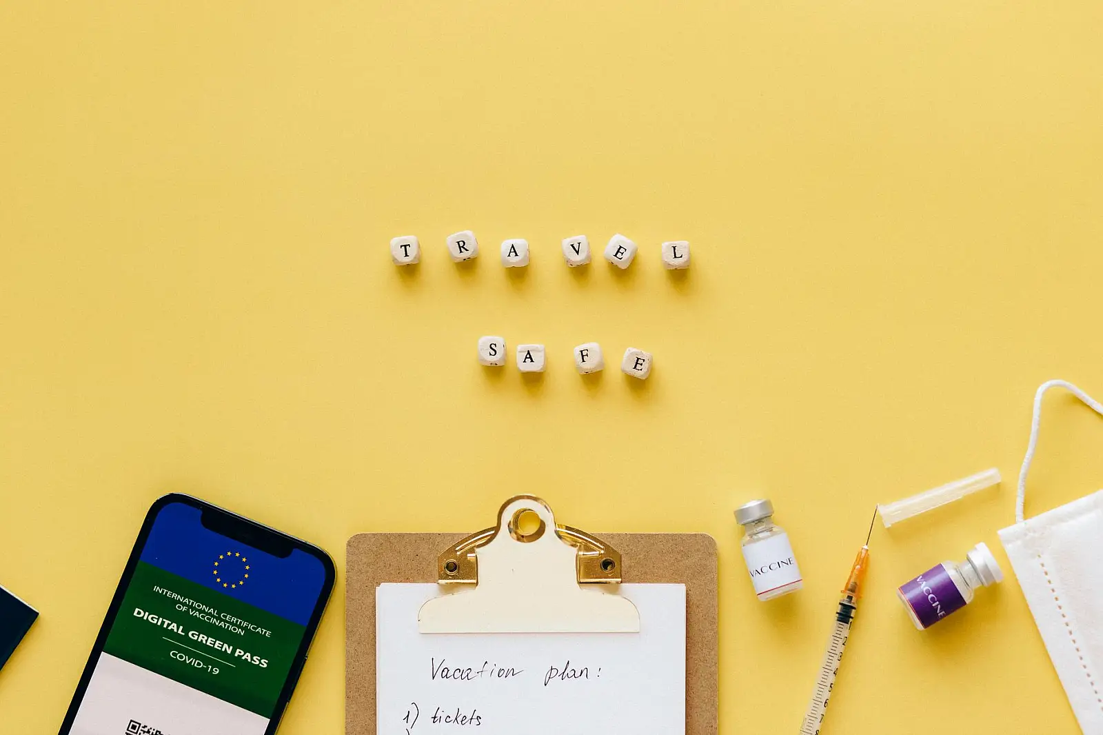 Flat lay showing travel safety essentials like a digital green pass, vaccine, and mask.