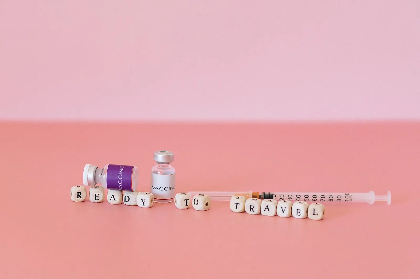 Close-up of vaccine vials, syringe, and the words 'Ready to Travel' on a pink background.
